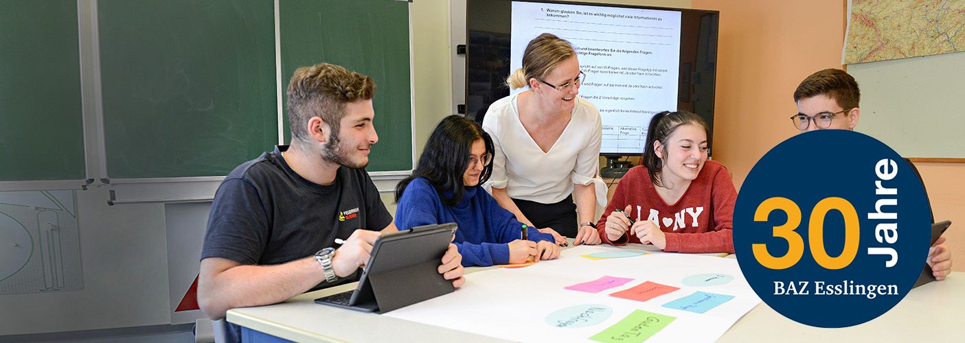 Vier Personen sitzen an einem Tisch, auf dem große Blätter in unterschiedichen Farben liegen und werden von einer fünften Person unterrichtet, die zwischen ihnen steht. Im Hintergrund ist eine Tafel und ein großer Bildschirm mit Informationen zu sehen. 