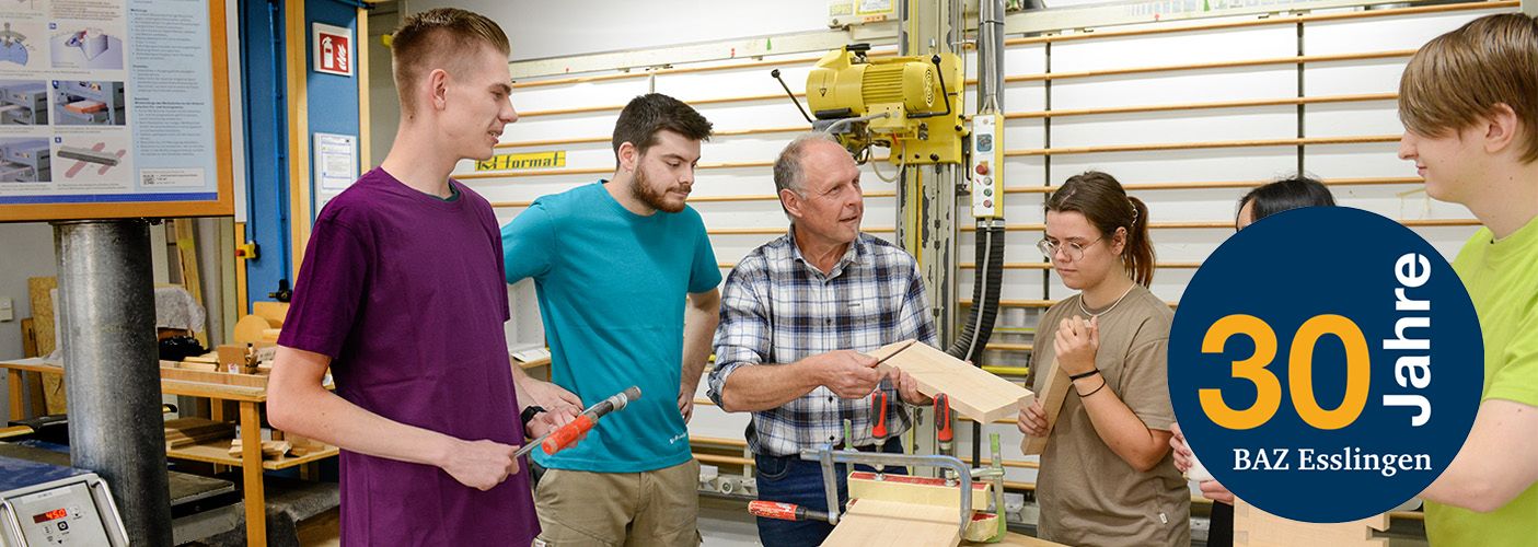 Ein Ausbilder in einer Werkstatt informiert fünf jungen Personen, die links und rechts neben ihm stehen, über die Bearbeitung von Holz. 